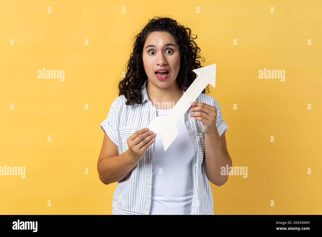 Portrait of amazed excited woman with dark wavy hair standing holding white arrow pointing aside, looking at camera with open mouth. Indoor studio shot isolated on yellow background. Stock Photo
