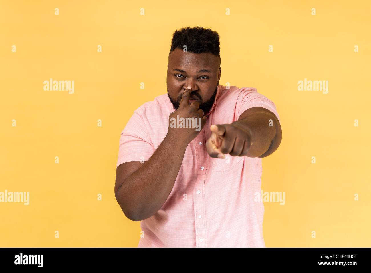 You are liar. Portrait of man wearing pink shirt standing with finger on her nose and showing lie gesture, pointing to camera. Indoor studio shot isolated on yellow background. Stock Photo