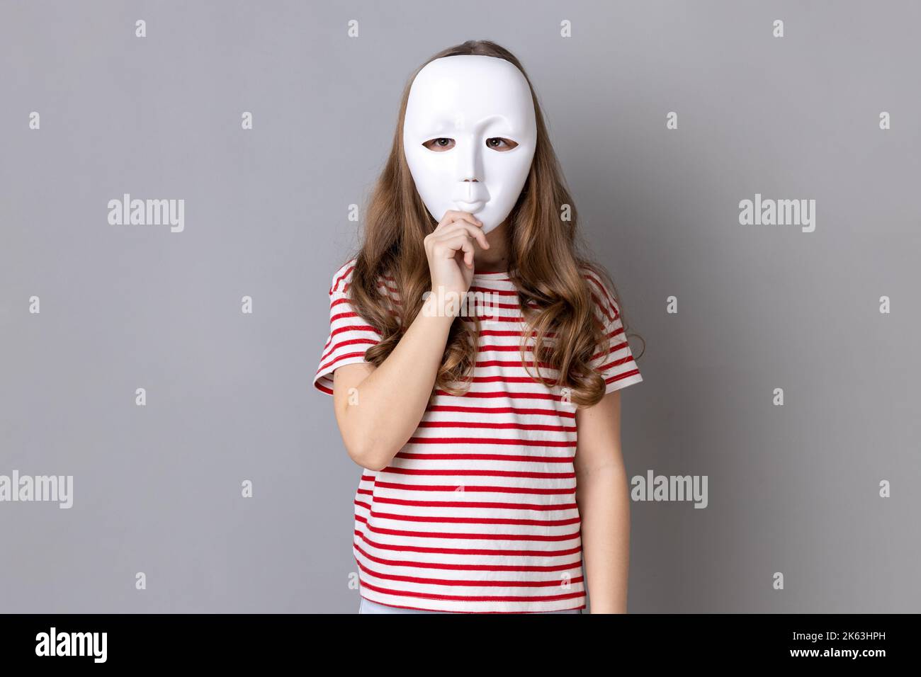 Portrait of anonymous unknown little girl wearing striped T-shirt covering her face with white mask, hiding her real personality, anonymity. Indoor studio shot isolated on gray background. Stock Photo