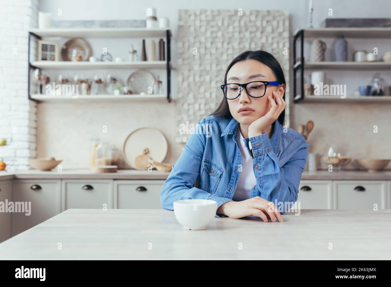sad and thinking woman in kitchen, depressed asian woman sitting at table at home, young wife in glasses disappointed. Stock Photo