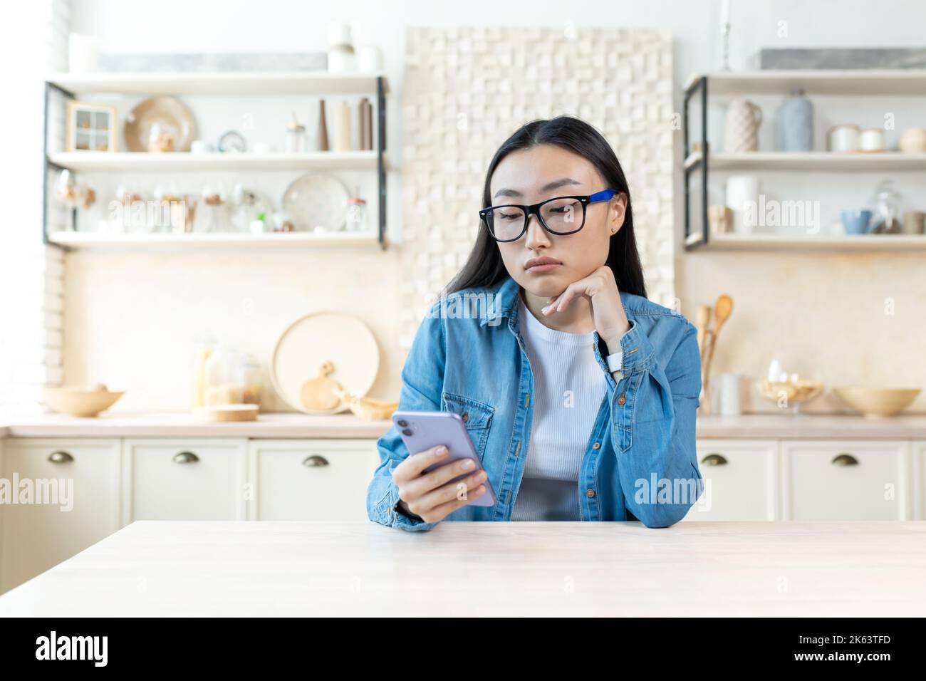 Sad woman at home sitting in kitchen reading news on phone, depressed asian woman holding smartphone in glasses and shirt. Stock Photo