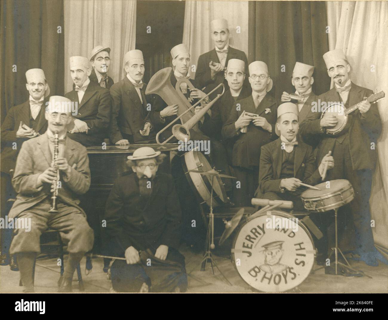 A band, most of whom are wearing disguise of heavy eyebrows and moustaches, presumably a gimmick during a performance, or just for the hell of it?! Somebody appears to be holding a cardboard tuba, and many of them are holding harmonicas so perhaps this is a comedy amateur performance? B'hoys was a slang term for boys, particularly used during the First World War. Stock Photo