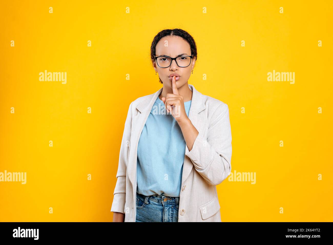 Secret concept.Pretty, cautious hispanic or brazilian young business woman with glasses, holds finger near lips, saying hush be quiet, shhh gesture, stand on isolated orange background,looks at camera Stock Photo