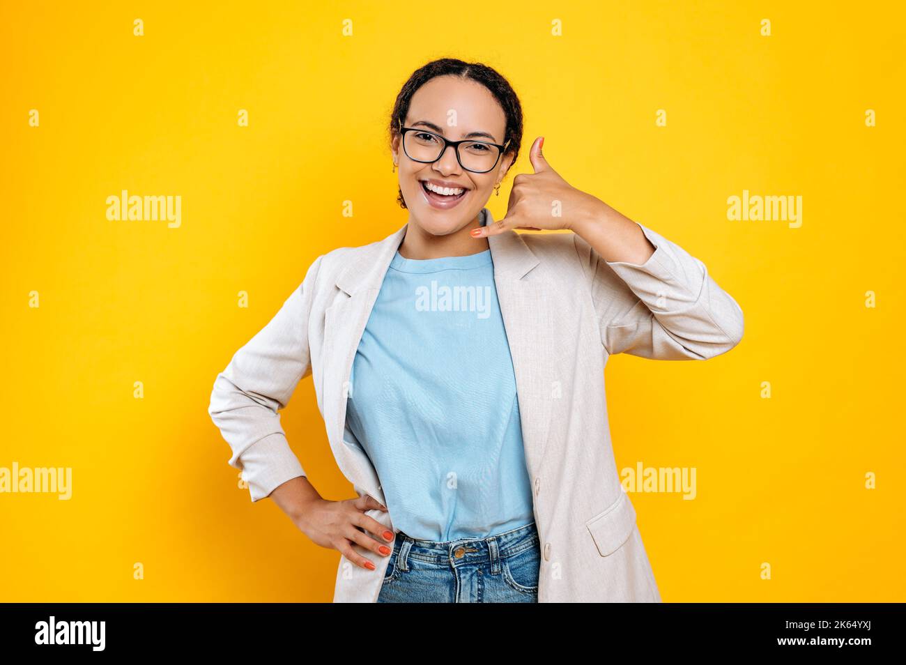 Cheerful friendly pretty hispanic or brazilian young woman, stylishly dressed, with glasses, doing phone gesture with hand, like says call me back, smiles at camera,stand on isolated orange background Stock Photo