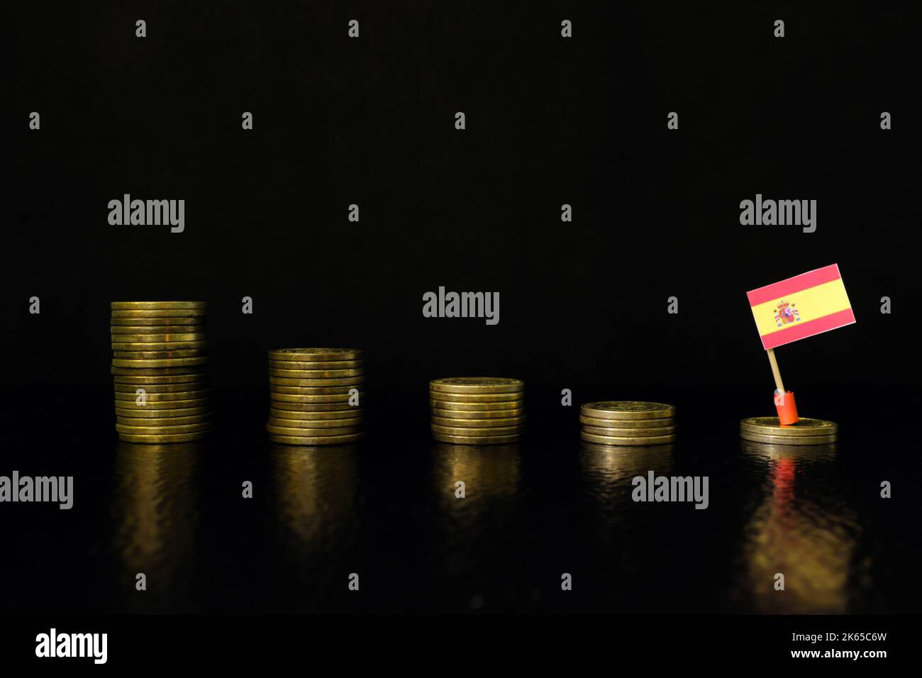 Spain economic recession, financial crisis and currency depreciation concept. Spanish flag in decreasing stack of coins in dark black background. Stock Photo