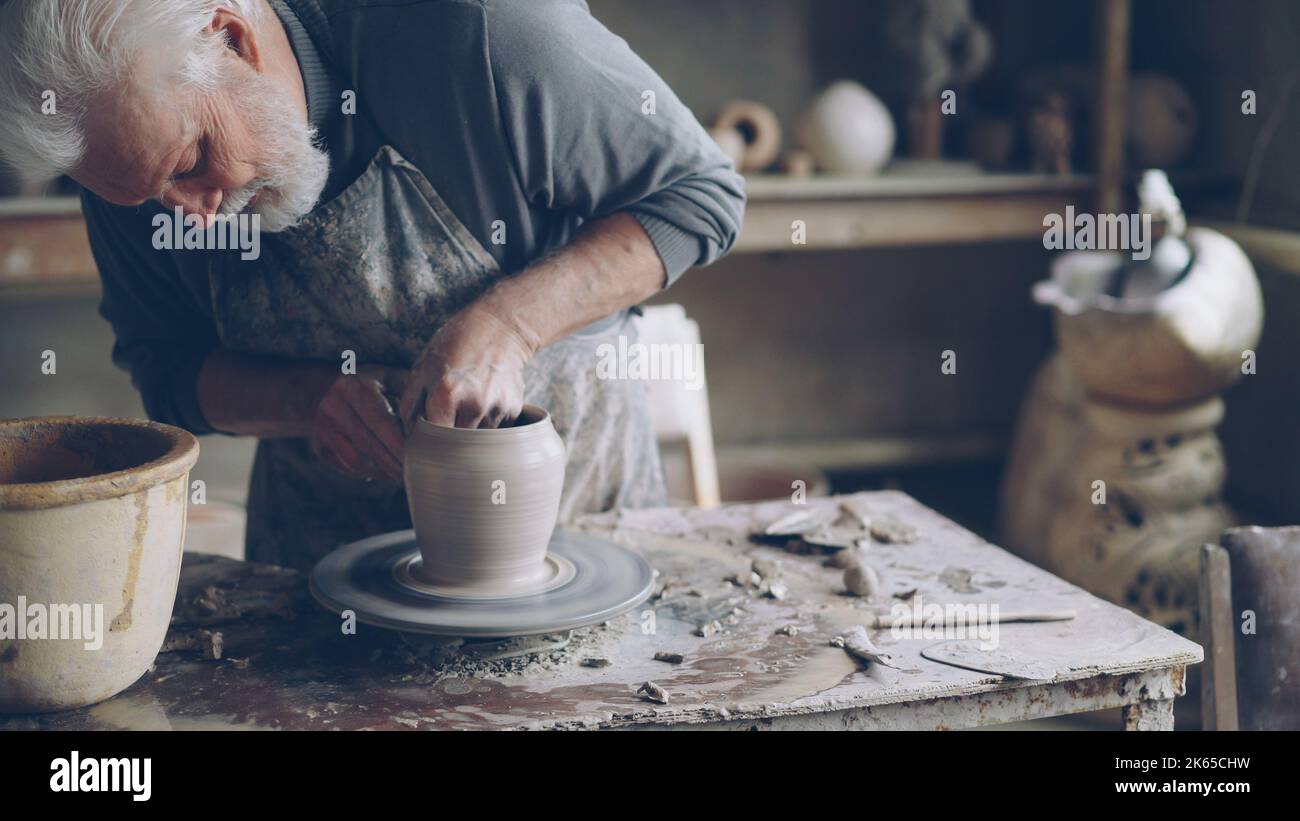 Experienced ceramist grey-haired bearded man is smoothing molded ceramic pot with wet sponge. Spinning throwing wheel, muddy work table and handmade clayware are visible. Stock Photo