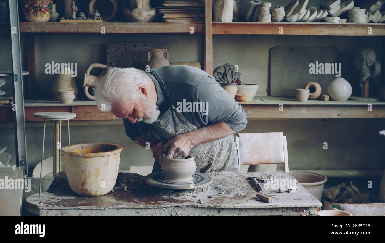 Skilled elderly male potter is producing beautiful ceramic pot on turning wheel in workplace