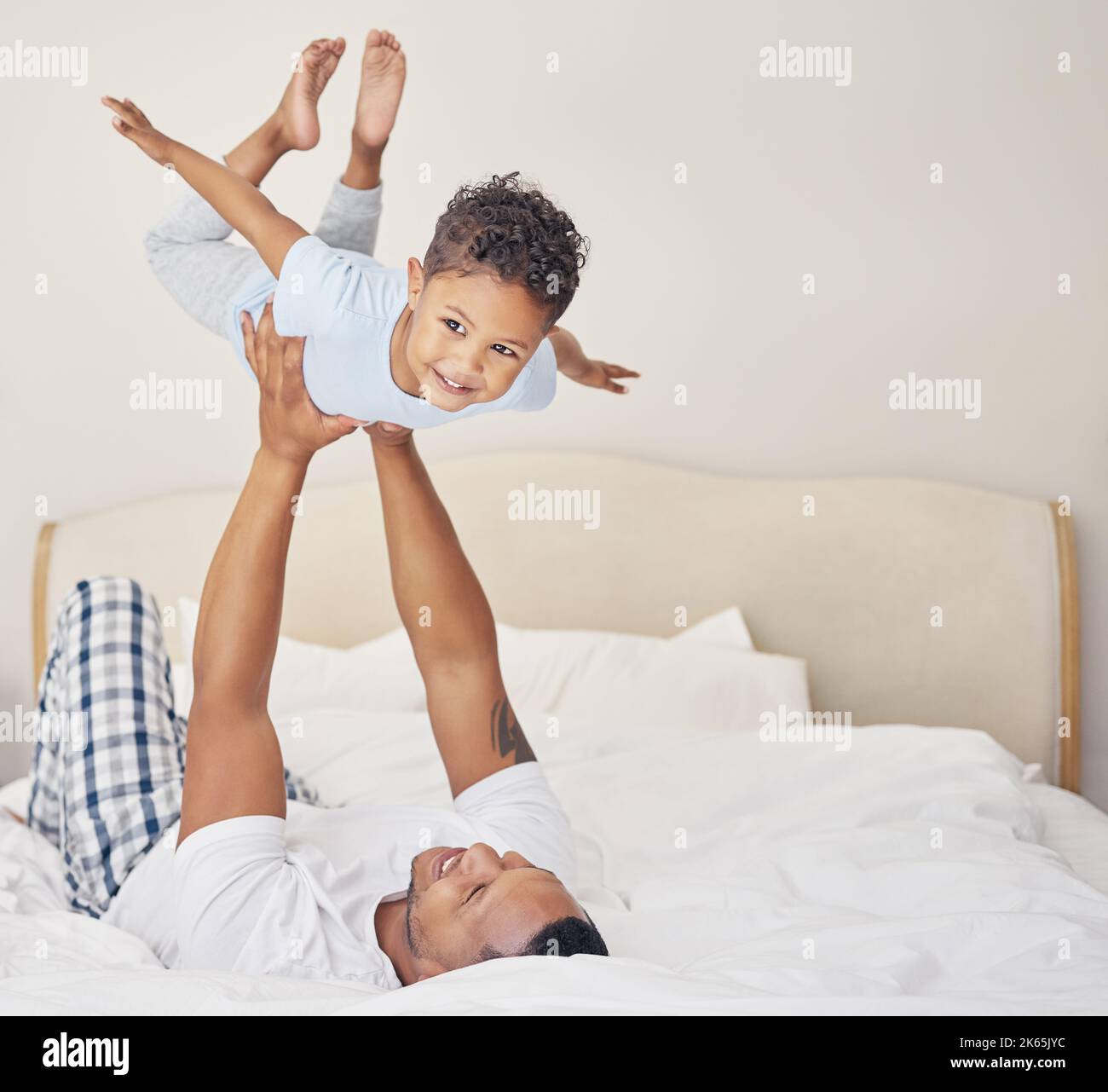 Joyful young single father lying on bed, lifting excited happy little child son at home. Little boy pretending to fly while having fun in bedroom with Stock Photo