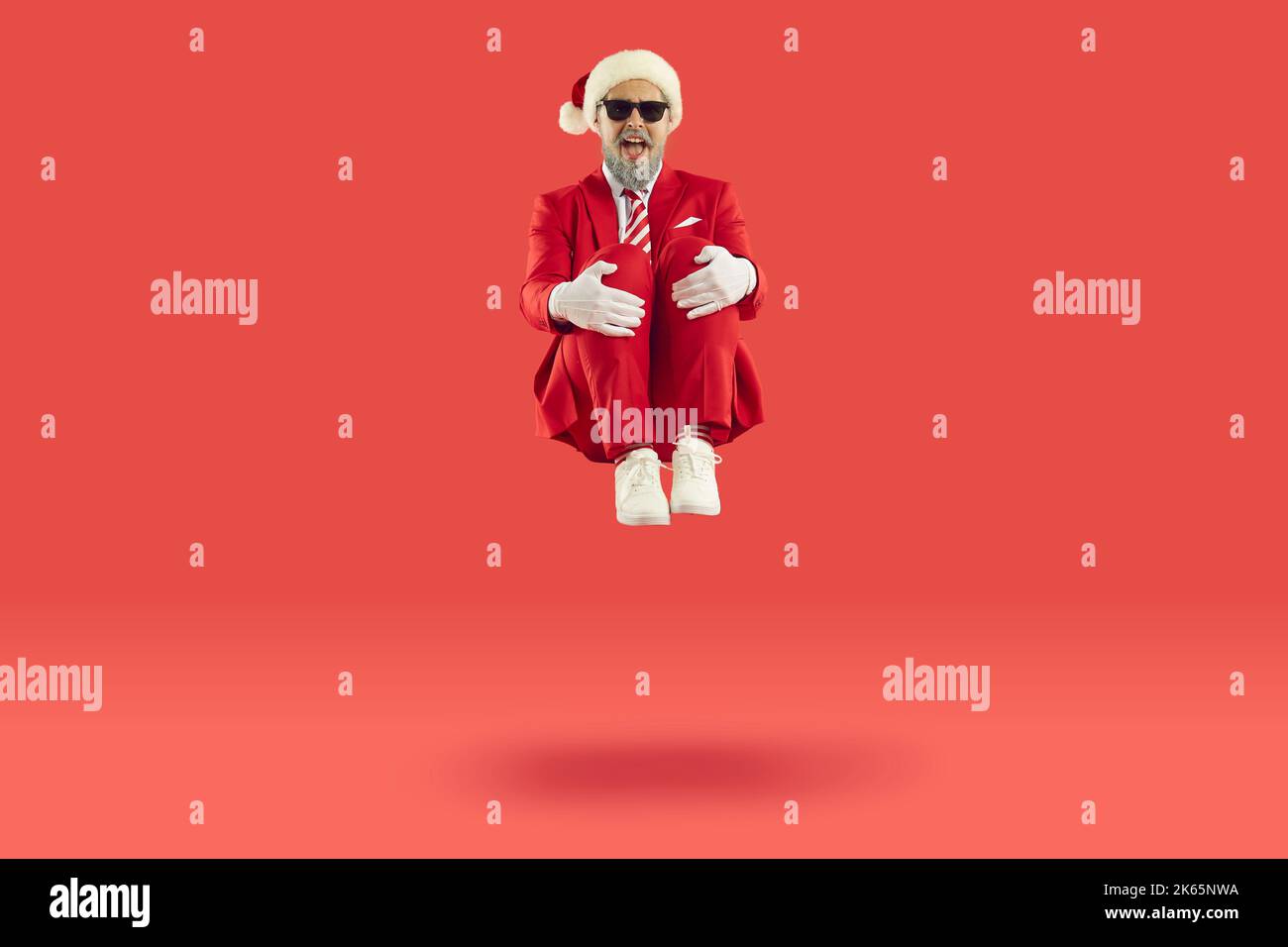 Old cheerful grey-haired man wearing Santa style is sitting levitating in air on red background. Stock Photo