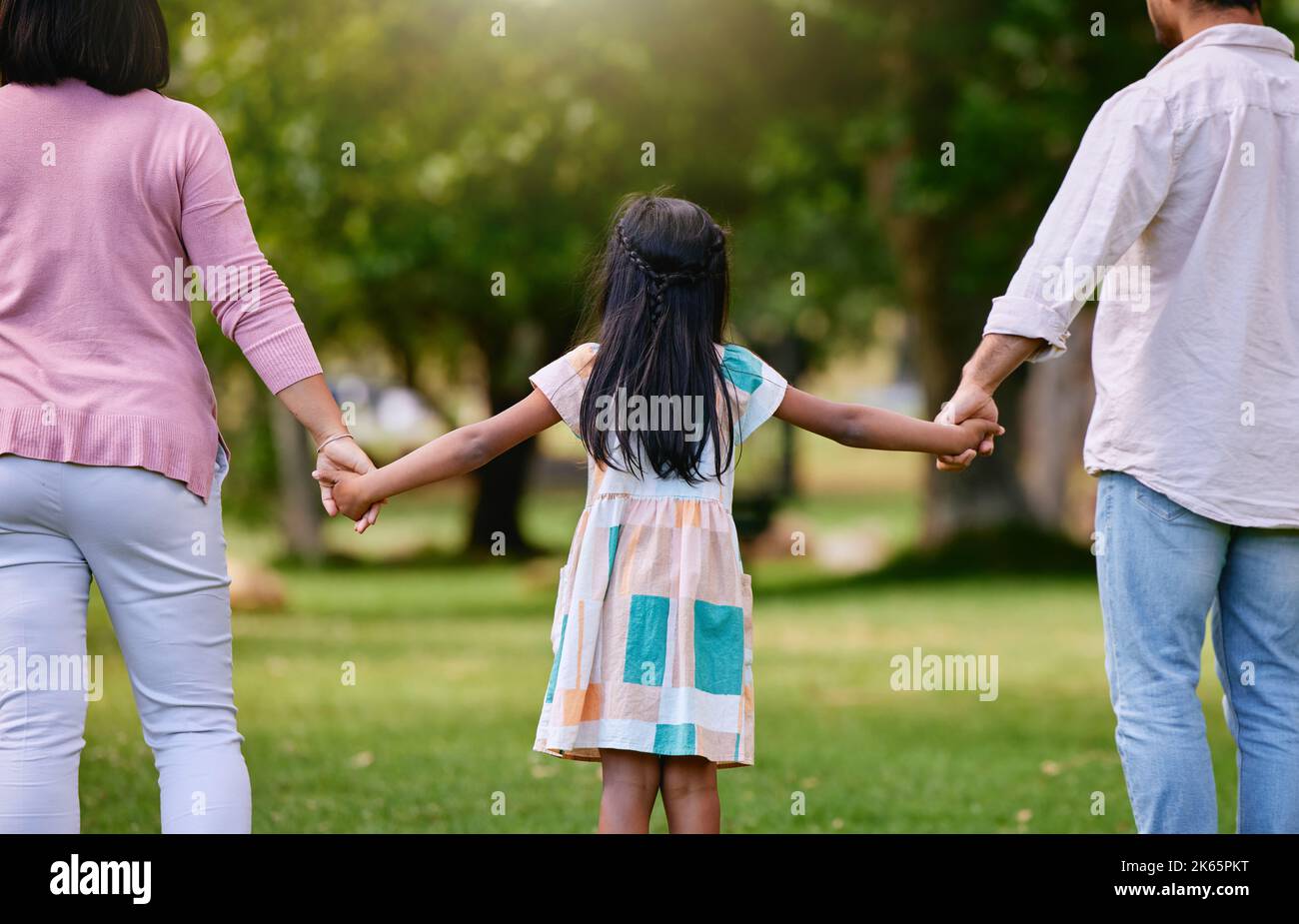 Rear view of parents and little girl holding hands while walking outdoors in park on a sunny day. Loving and caring family with one child bonding and Stock Photo