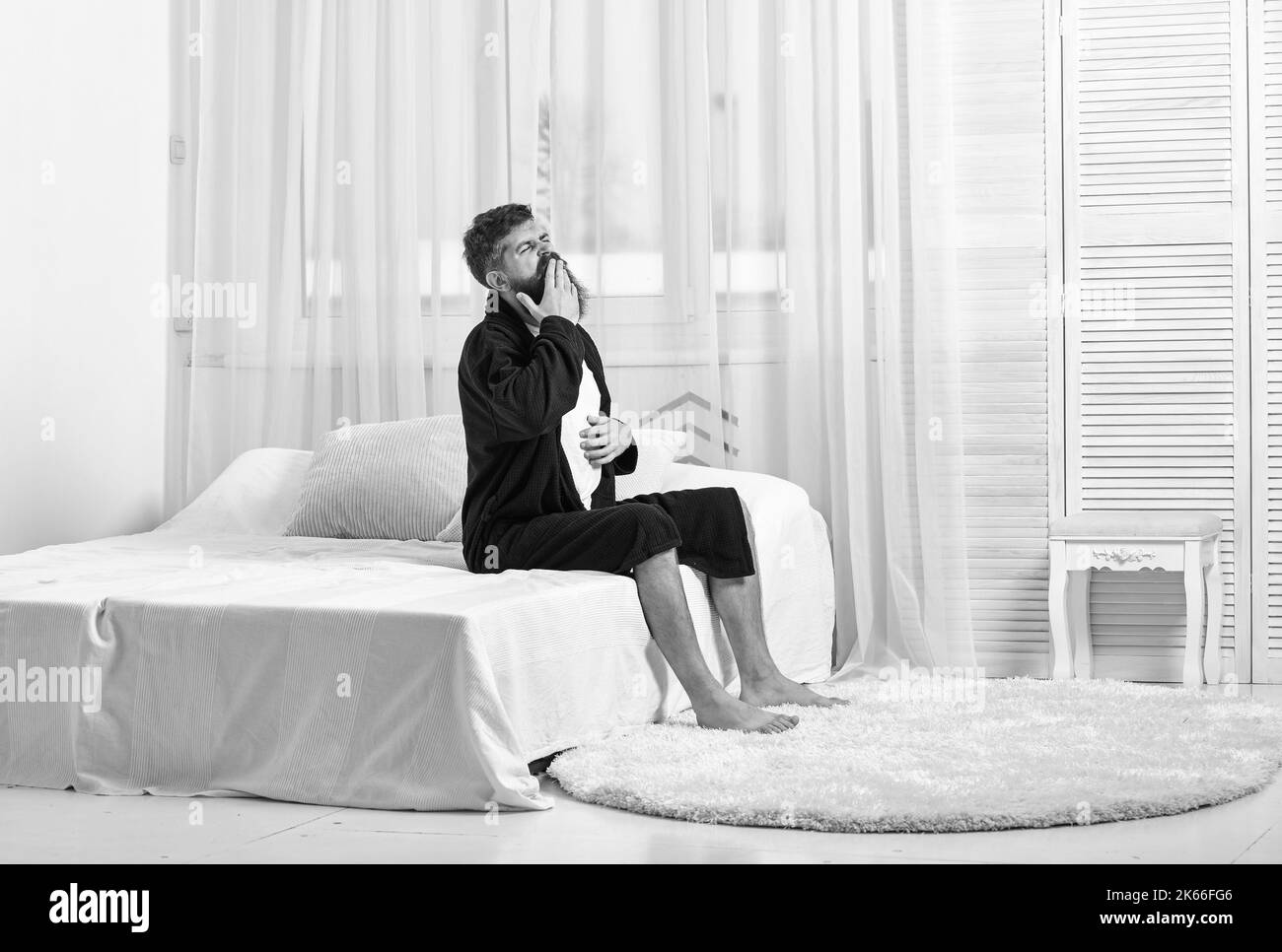 Awakening concept. Man in robe sits on bed, white curtains on ...