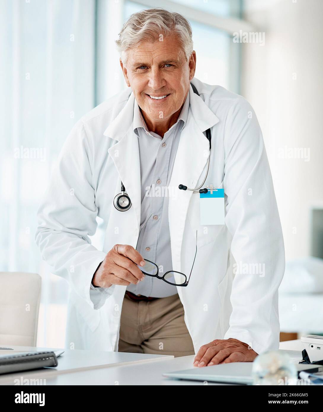 Experienced mature caucasian man doctor smiling and standing and holding his glasses in a hospital. One optimistic senior male general practitioner Stock Photo