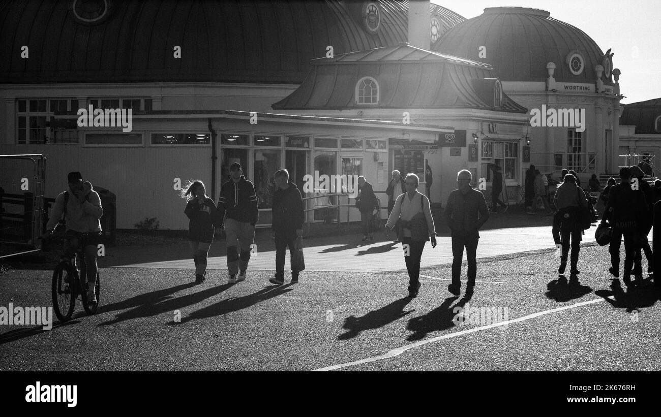 People walk in late afternoon sunshine on Worthing seafront next to the pier, West Sussex, UK. Stock Photo