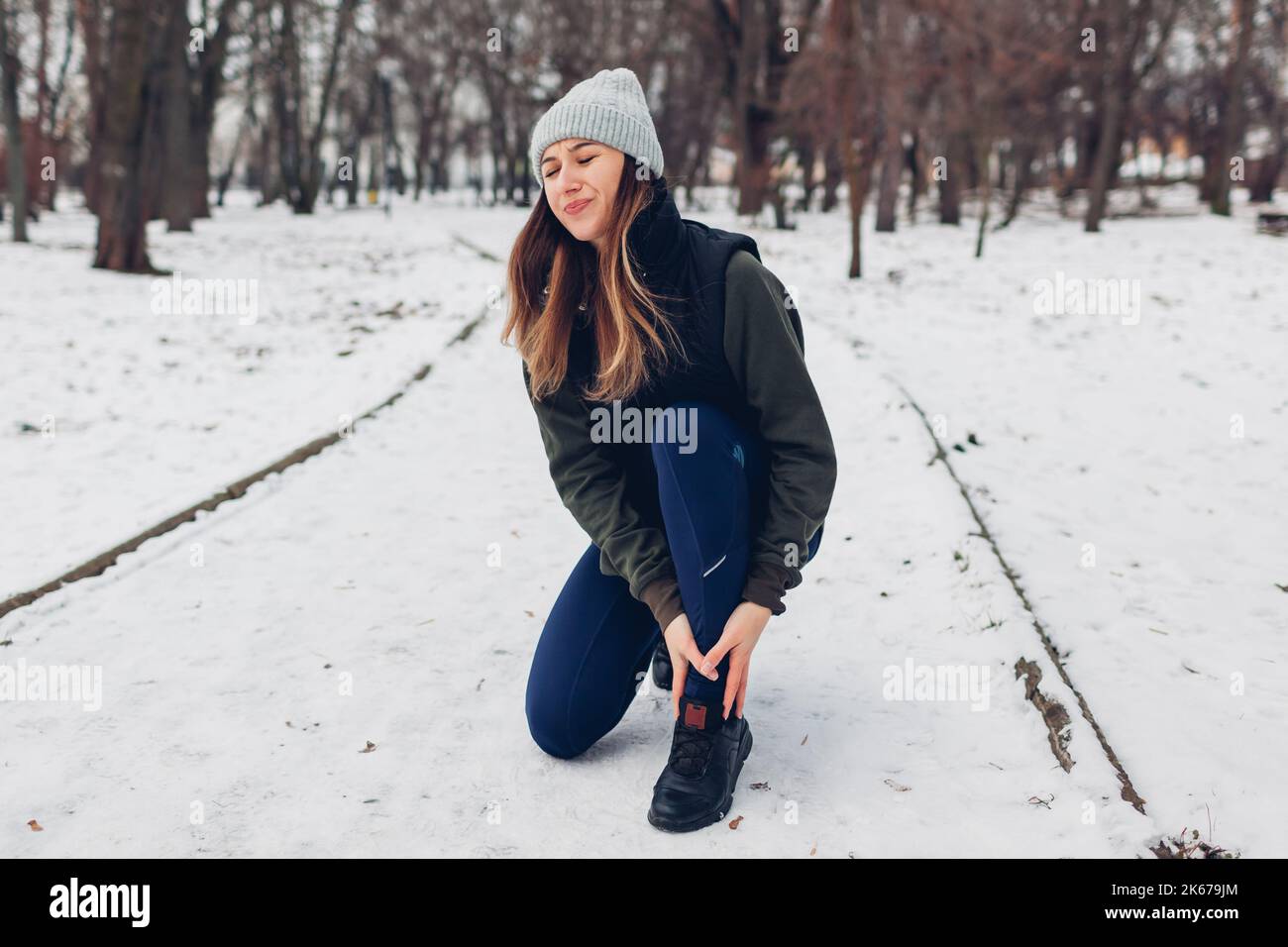 Runner injured leg during training in snowy winter park. Woman feels hurt, ankle pain. Dangerous exercises outdoors during cold weather Stock Photo
