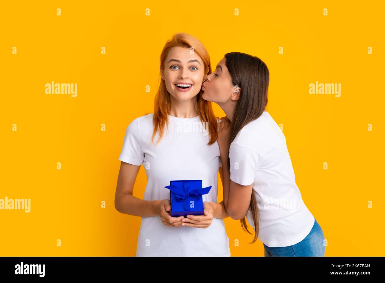 Child kissing mom. Mother and daughter with present gift. Teenager girl giving gift box to her mother on yellow background. Stock Photo