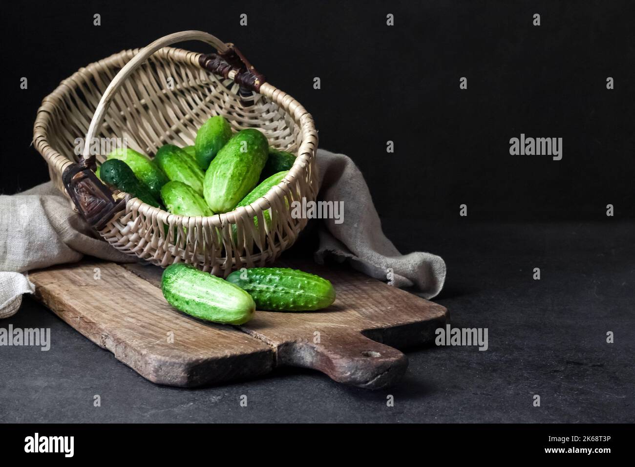 Fresh cucumbers in wicker basket on wooden board on black background. Stock Photo