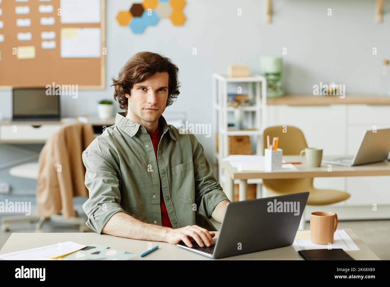 Portrait of long haired young man at office workplace using laptop and looking at camera with smile, IT professional, copy space Stock Photo