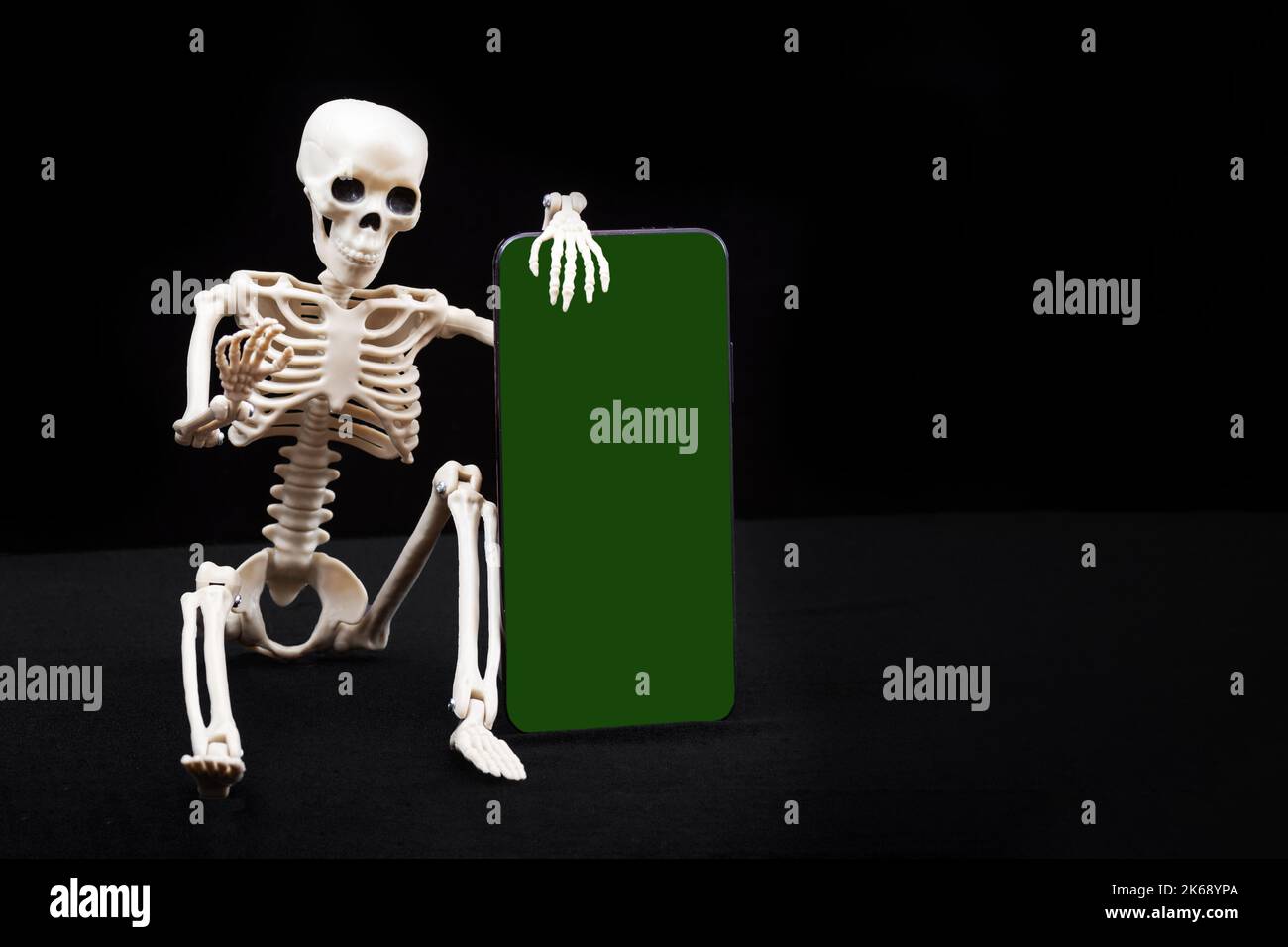A seated skeleton is holding a smartphone with a green chromakey screen isolated on a black background, mockup, copy space. Halloween online. Online S Stock Photo