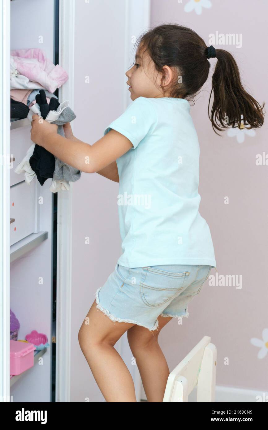 Beautiful little girl with long black hair brings wardrobe order, puts everything in its place in closet. Wardrobe with child's clothing. Stock Photo
