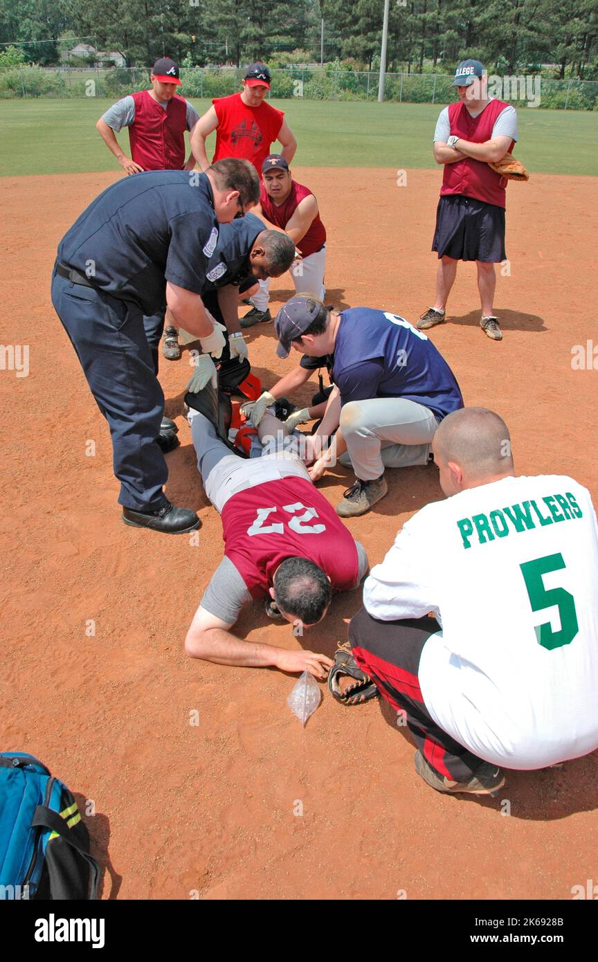 softball Player with broken leg is attended to by fire personal and wife as team members look on while laying on ball field during softball game Stock Photo