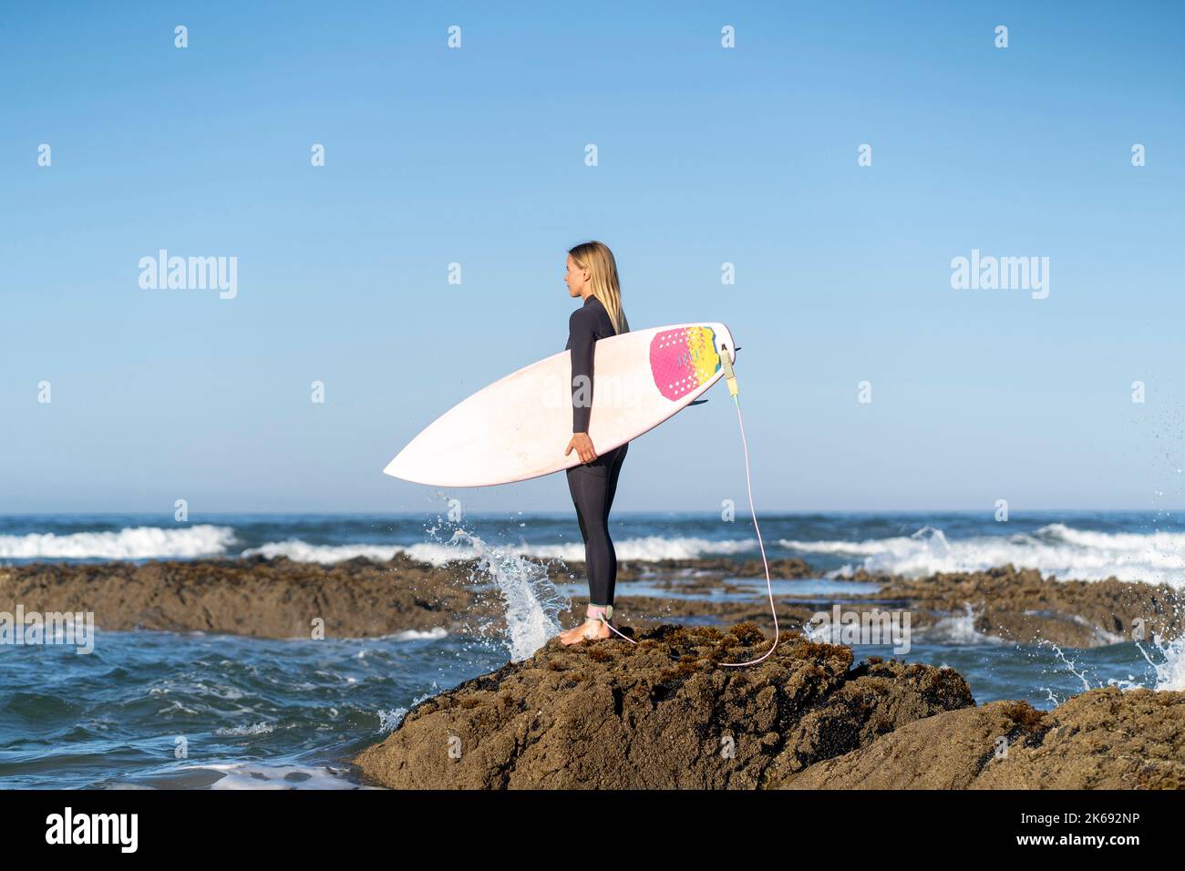 Surfer girl at the beach standing on a rock with her surfboard in the