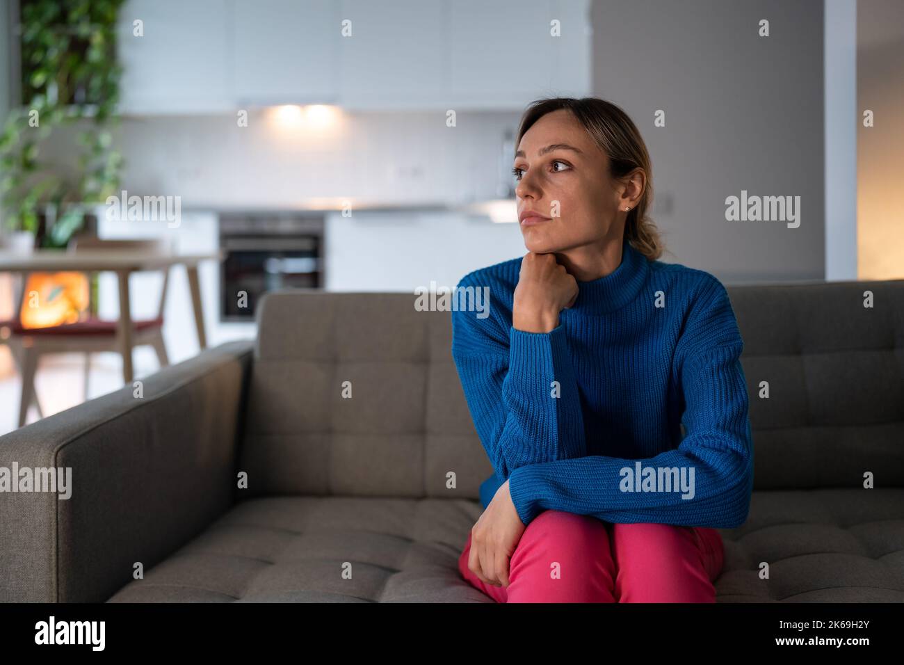 Pensive woman sits on comfortable sofa with exhausted and depressed expression in apartment Stock Photo