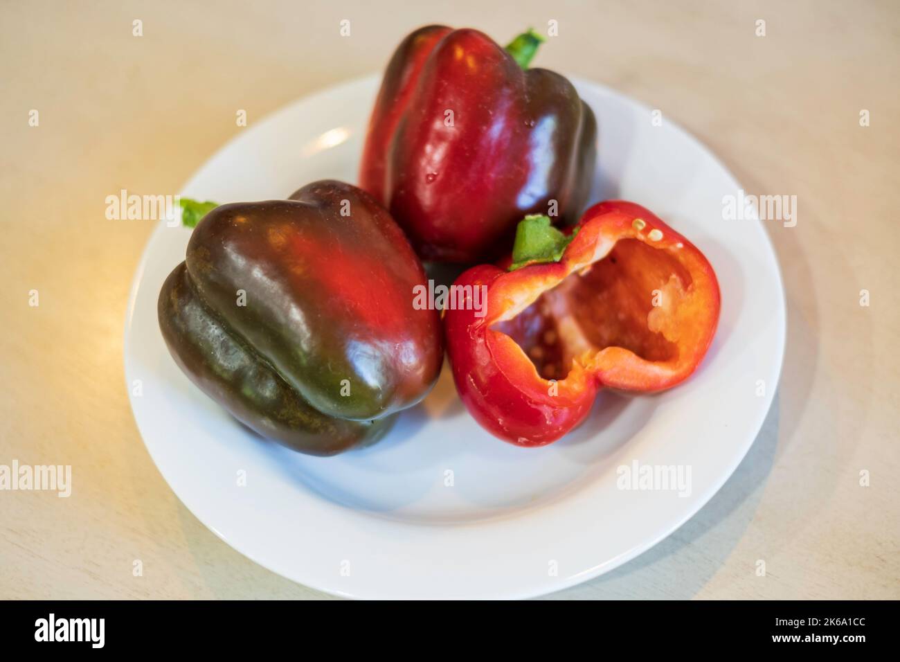 Three home grown red bell peppers, just turning red with one cut in half on a white plate. USA. Stock Photo