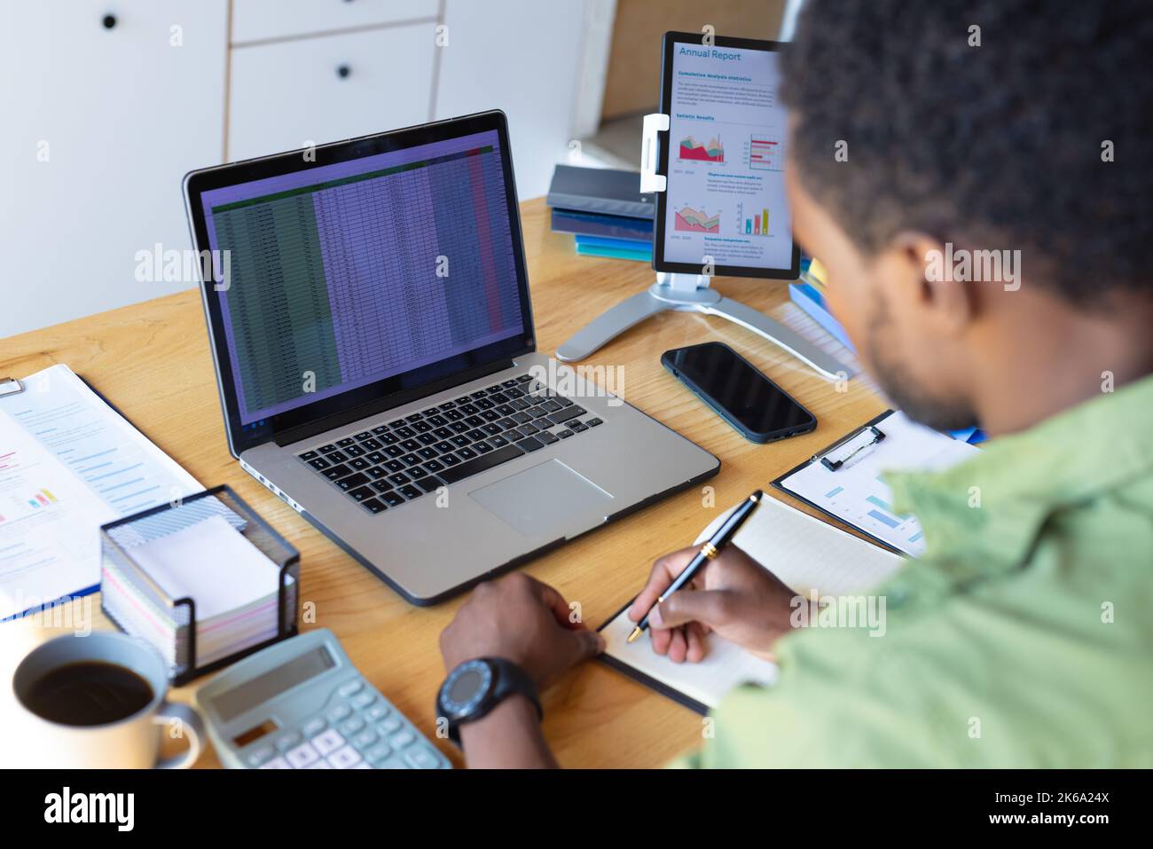 Happy african american man standing at table in kitchen, using laptop and taking notes Stock Photo