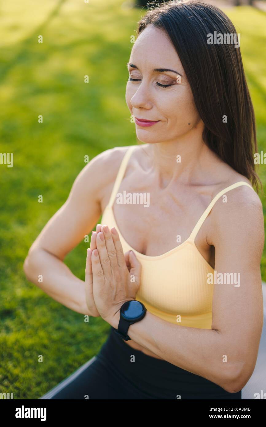 Beautiful brunette woman meditating in prayer position at park, breathing fresh air in the morning. Young female athlete keeping balance in yoga Stock Photo