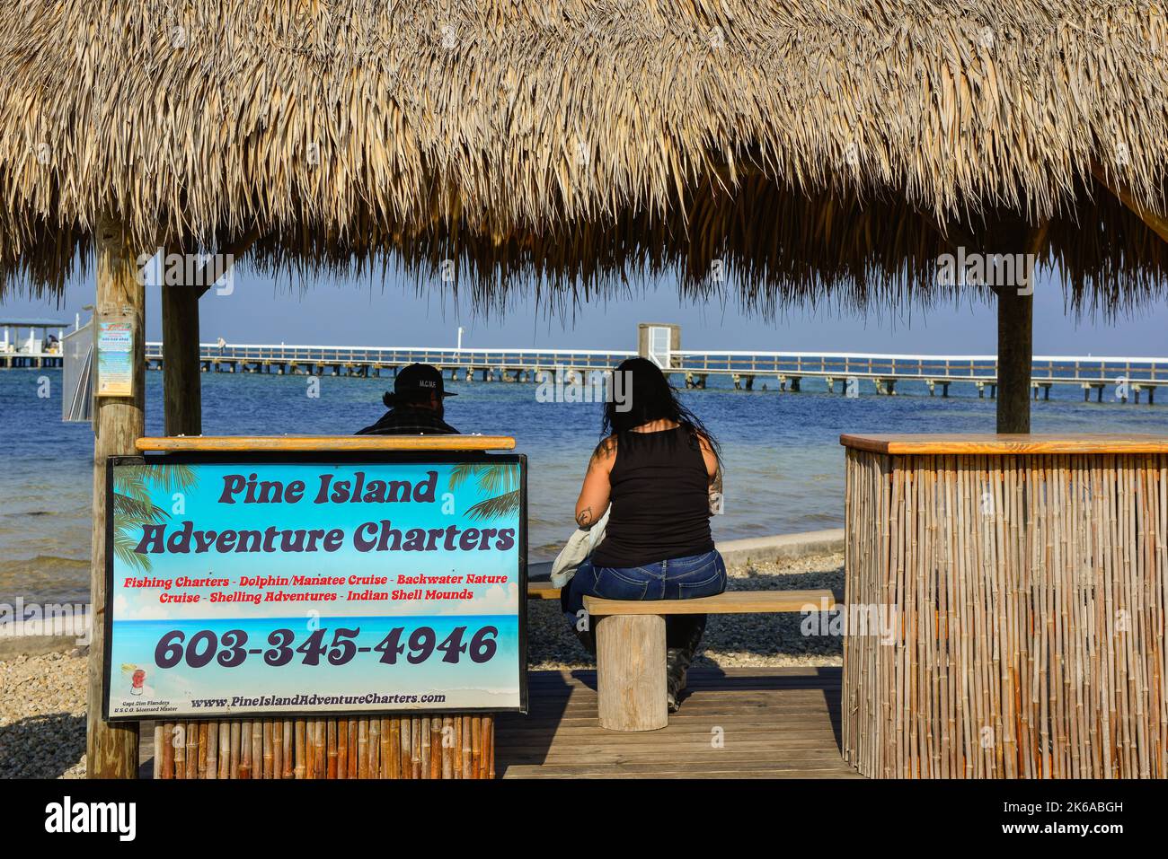 Rear view of two people under the shade of a thatched roof kiosk for Adventure Charter tours in Bokkelia, FL on the northern tip of Pine Island Stock Photo
