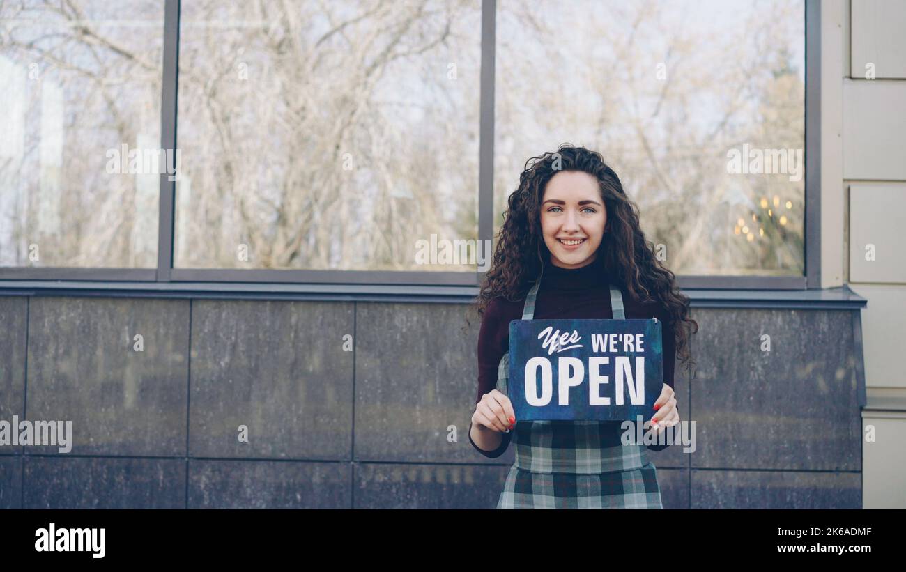 Portrait of cheerful pretty waitress in apron holding yes we are open sign standing outside opposite window and smiling looking at camera. Starting catering business concept. Stock Photo