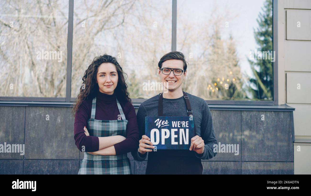 Portrait of attractive friends business partners opening restaurant and holding we are open sign in front of window outside building. Successful start-up concept. Stock Photo