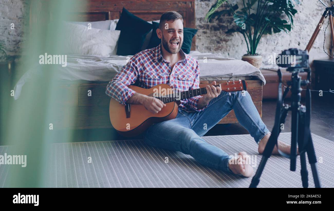 Young handsome musician popular blogger is recording video with camera playing the guitar and singing song sitting on floor in bedroom. Blogging and music concept. Stock Photo