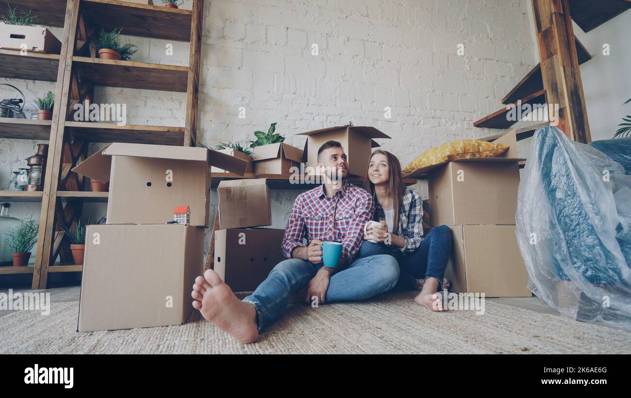 Adorable young couple is talking and holding mugs while sitting on floor of new flat after relocation. Numerous boxes, packed furniture and plants are visible. Stock Photo