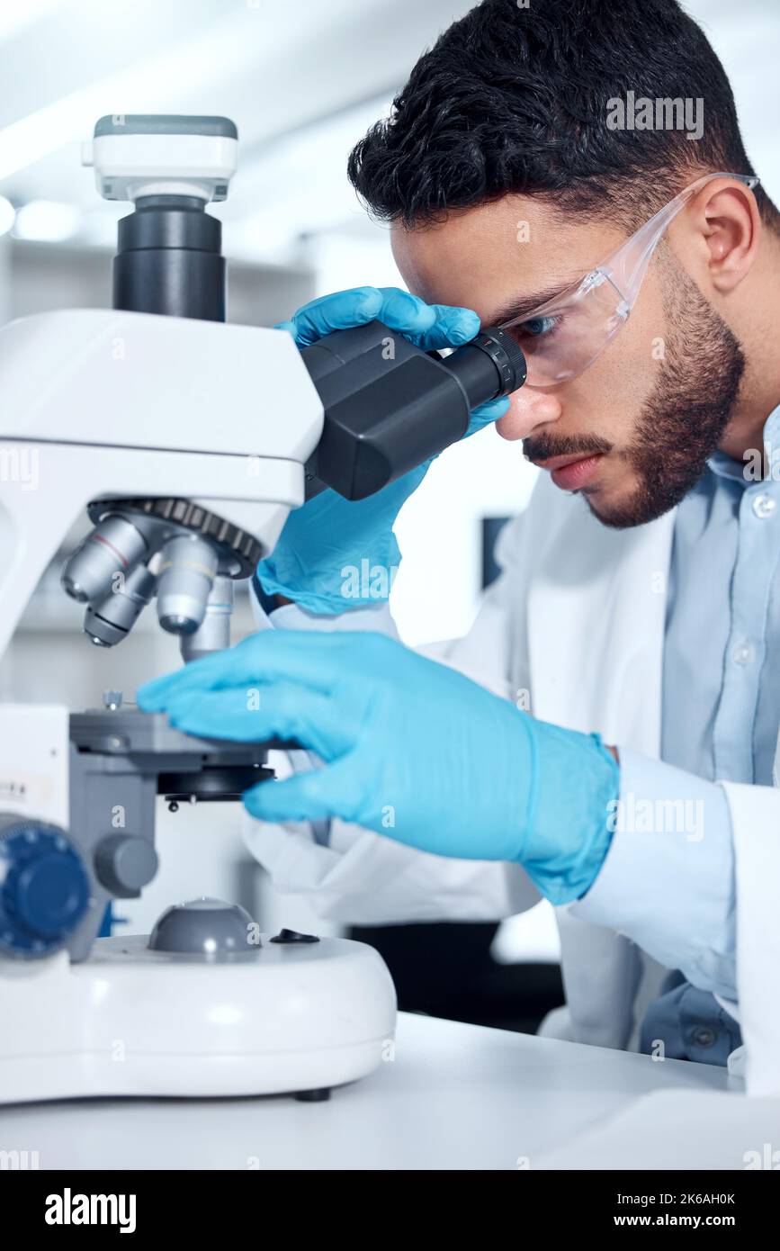 One mixed race scientist wearing safety goggles and gloves analysing medical test samples with a microscope in a lab. Young indian man developing a Stock Photo