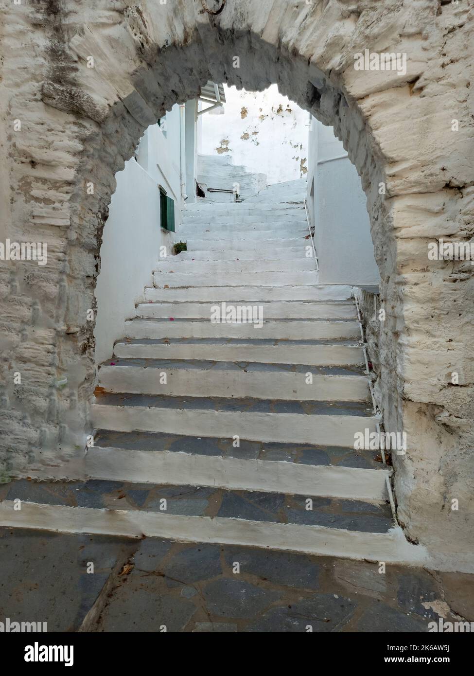 Greece, Tinos island, Cyclades. Arched stonewall entrance in front of stone paved stairs with white risers between whitewashed houses. Vertical Stock Photo