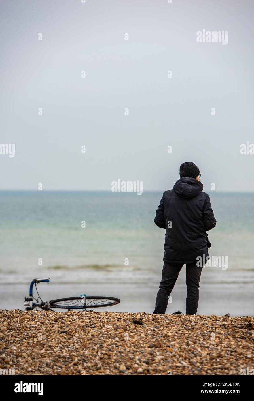 person standing alone on a beach next to a bicycle laying on the shingle looking out to sea daydreaming on a cold autumnal day. Stock Photo