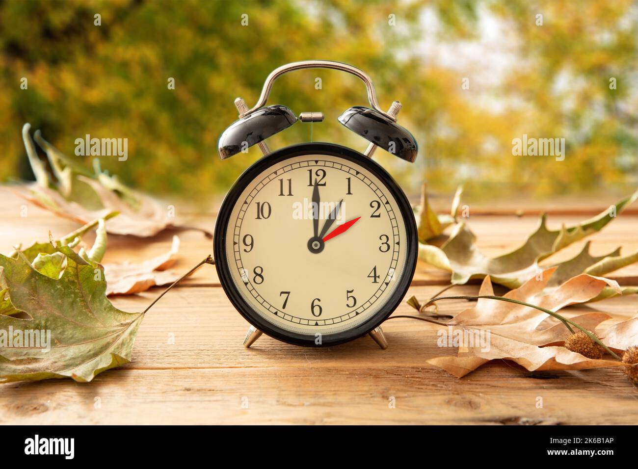 Fall Back one hour. Daylight Saving Time, Black alarm clock with time change on wooden table. Autumn trees and leaves background Stock Photo