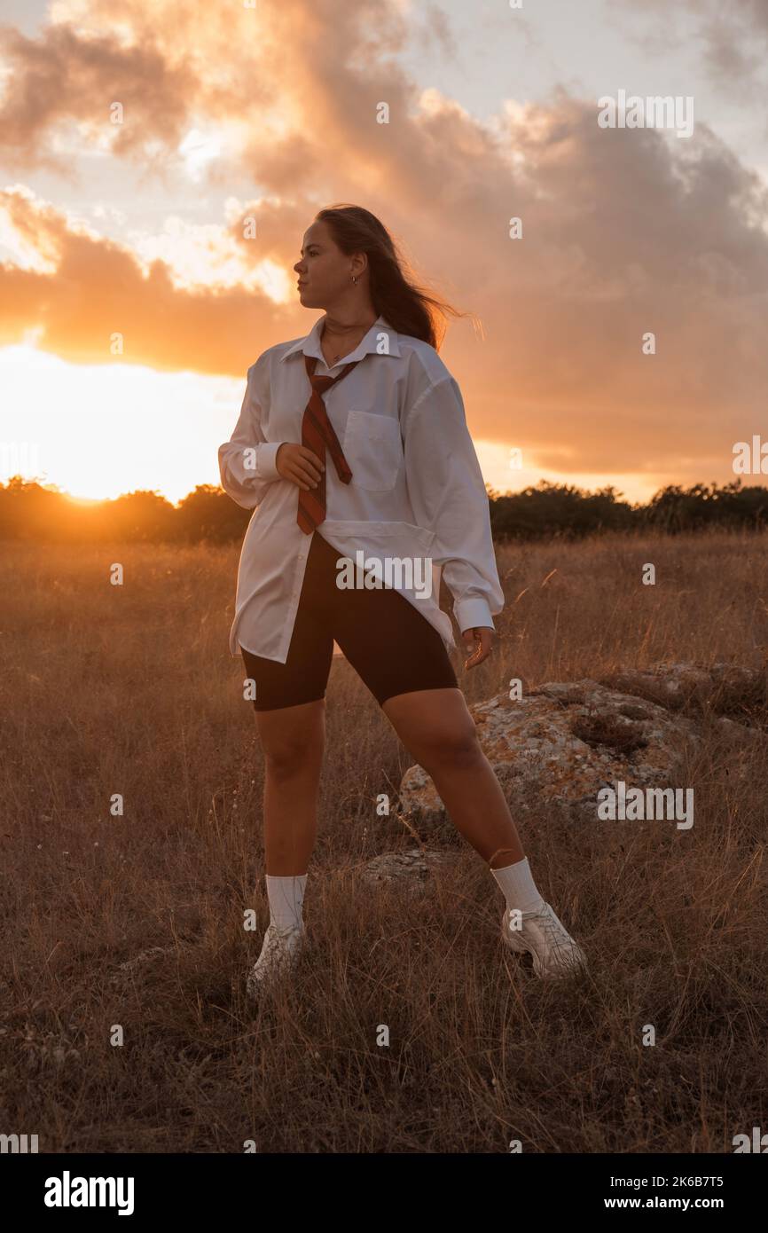 A white girl with black hair in a white long shirt, tie and black leggings stands and looks away in a field at sunset. A schoolgirl in a school uniform looks to a bright future. A fat girl is thinking about losing weight. High quality photo. Stock Photo