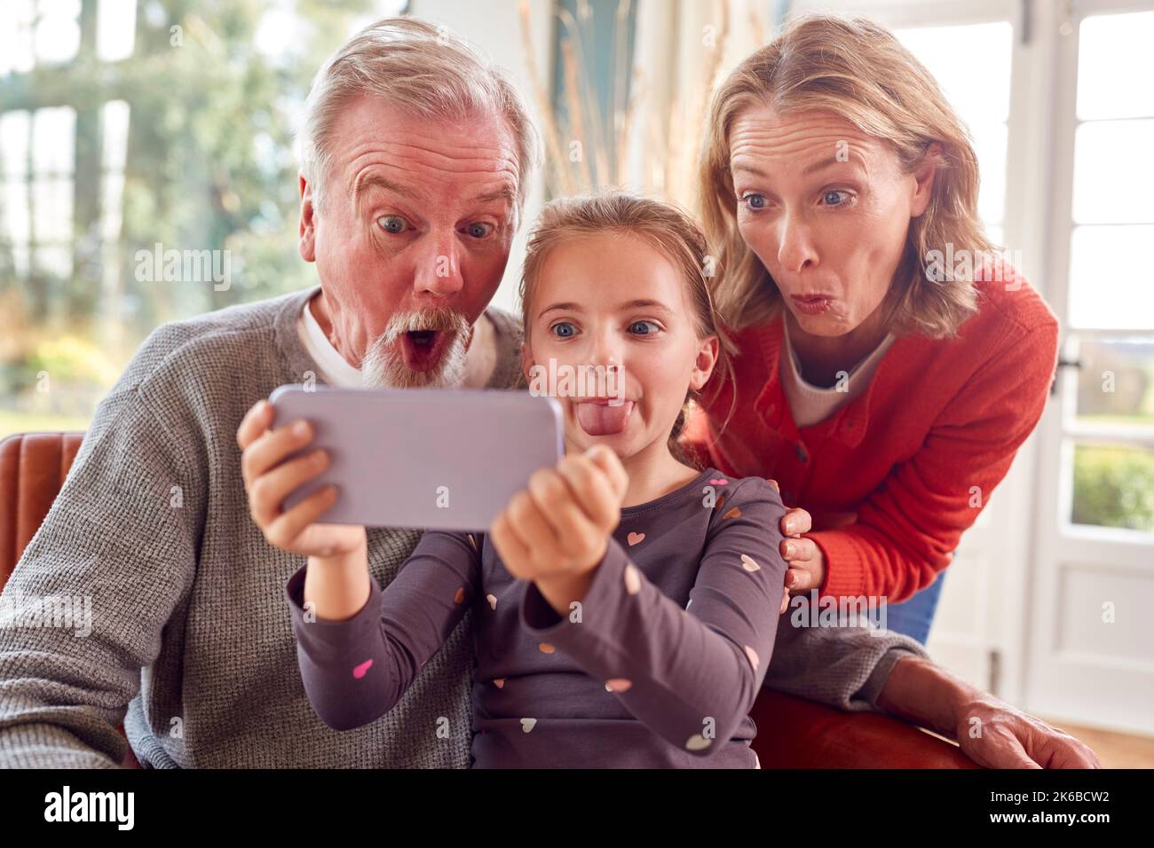 Grandparents With Granddaughter Pulling Faces Posing For Selfie On Mobile Phone At Home Together Stock Photo