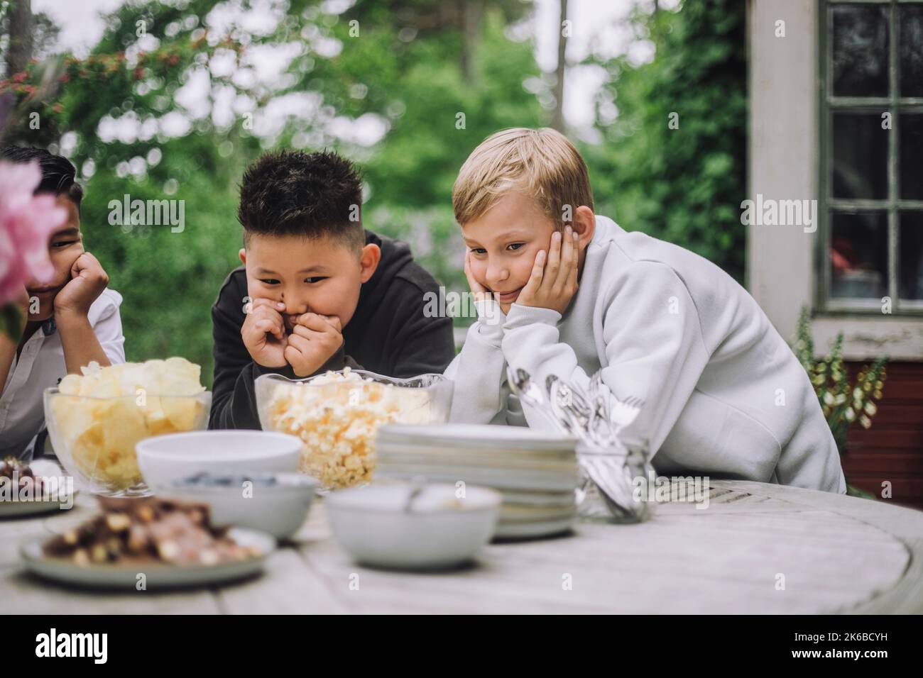 Curious boys leaning on elbows while looking at snacks bowl on table Stock Photo