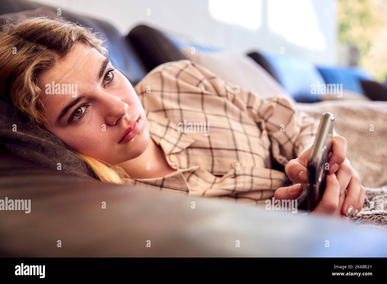 Unhappy Teenage Girl Lying On Sofa With Mobile Phone Being Bullied Online At Home Stock Photo