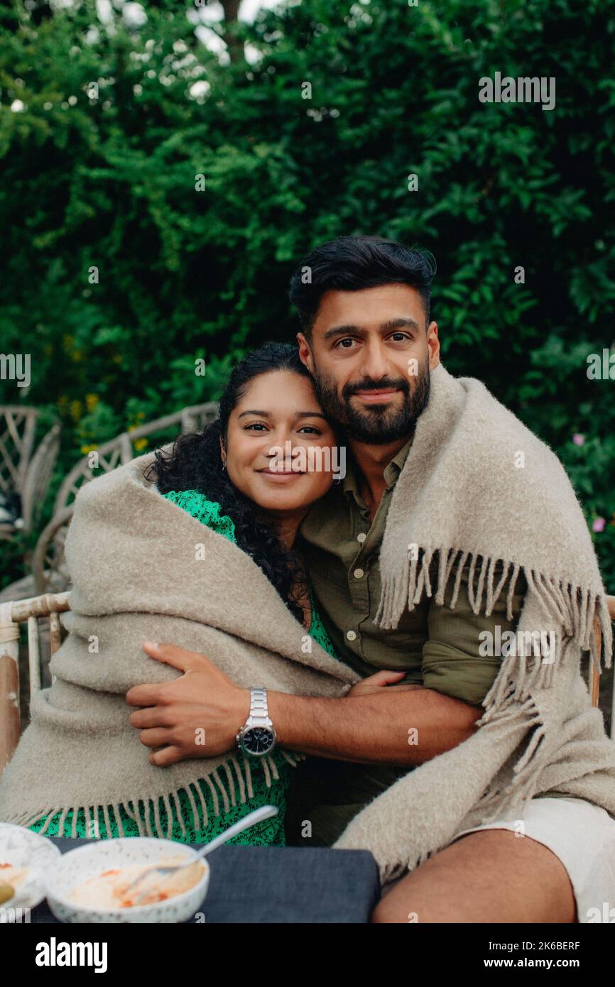 Portrait of man and woman covered in blanket embracing each other while sitting on bench Stock Photo