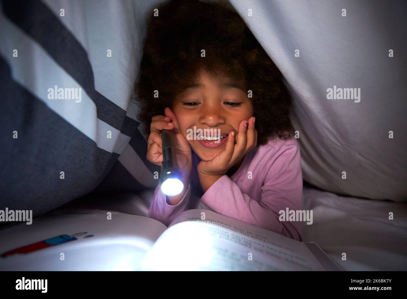 Girl Reading Book Under Duvet In Bed At Night Using Torch Stock Photo