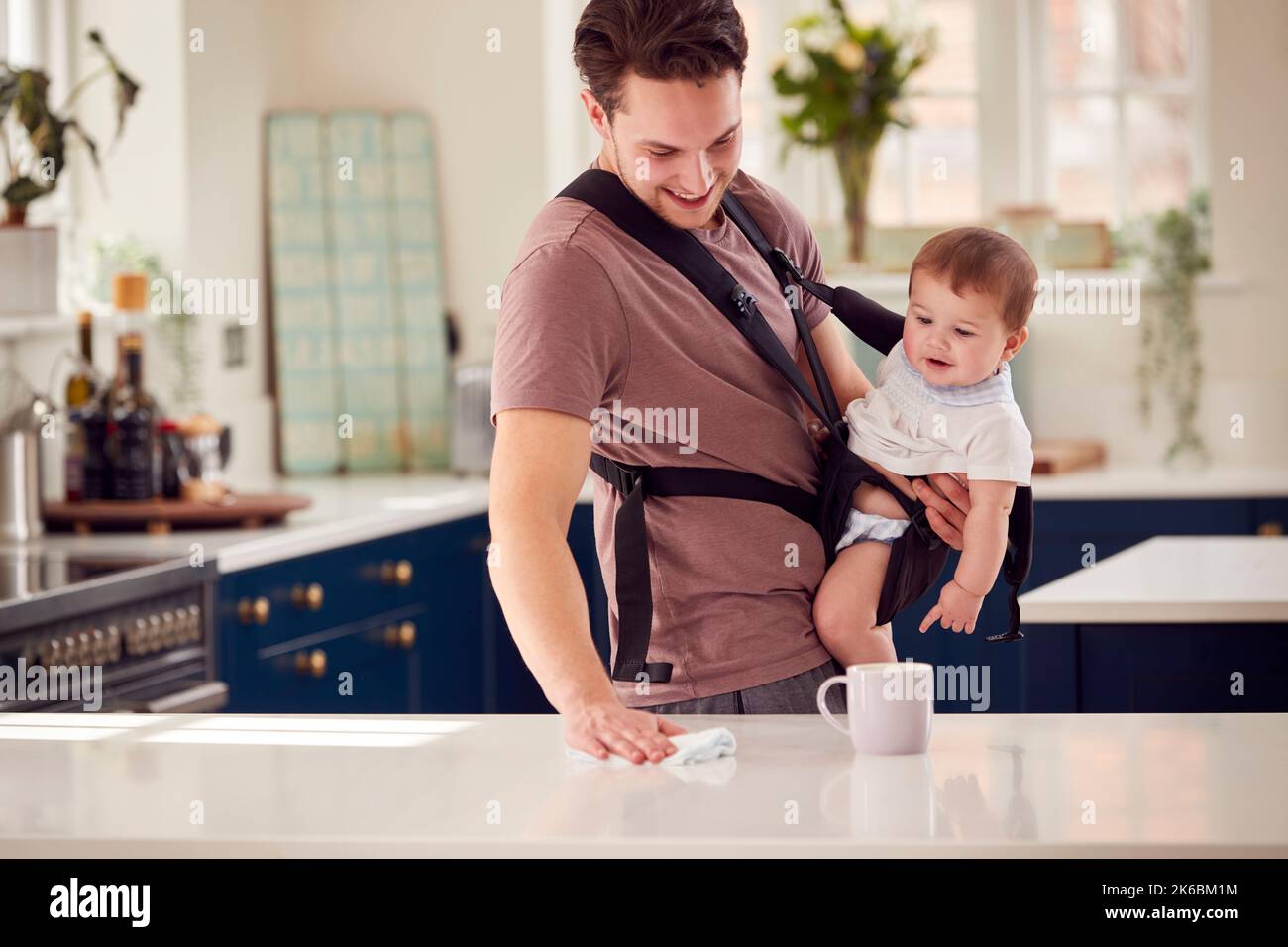 Transgender Father Cleaning Kitchen Looking After Baby Son In Sling Stock Photo