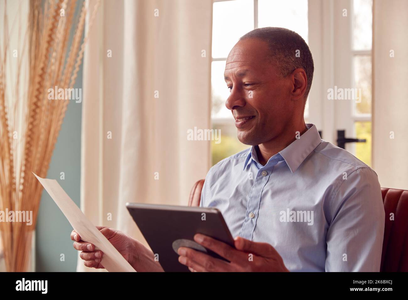 Mature Man Or Businessman Working From Home Sits In Armchair Reading Document Using Digital Tablet Stock Photo