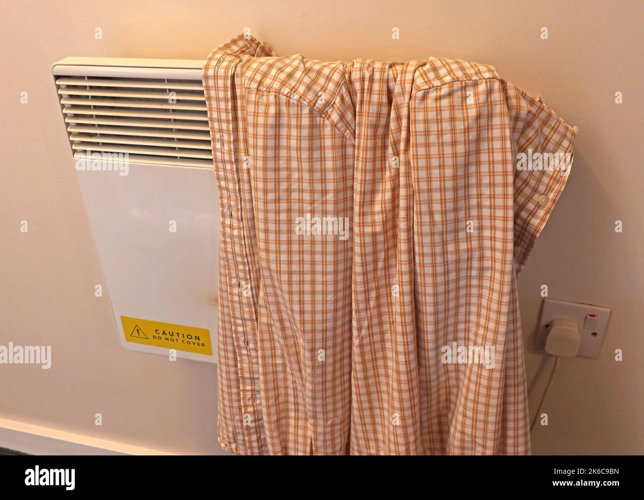 Wall mounted electric space heater, with shirt drying on the top, covering it. Sign 'caution do not cover' Stock Photo