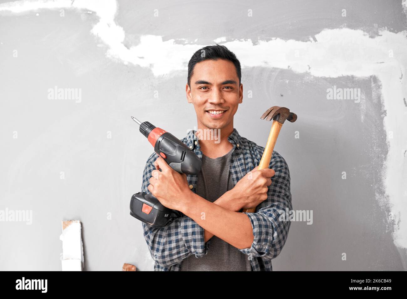A young Asian man smiles while holding drill and hammer for home DIY improvement Stock Photo