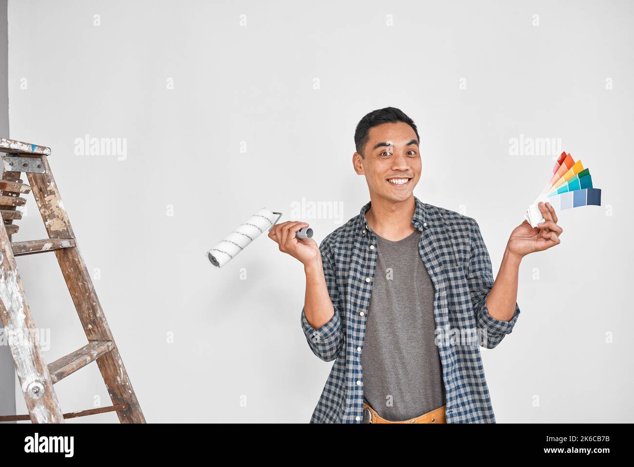 An attractive Asian man holds up paint roller and bright swatches for blank wall Stock Photo