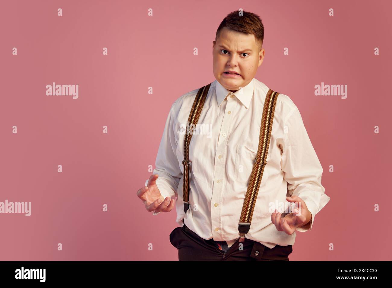 Anger. Studio shot of cute big obese teen boy in white shirt and shorts with suspenders isolated over pink background. Emotions, retro fashion Stock Photo